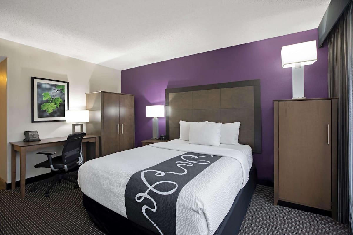A hotel room with a large bed, a desk with a chair, a wardrobe, and a nightstand with a lamp.
