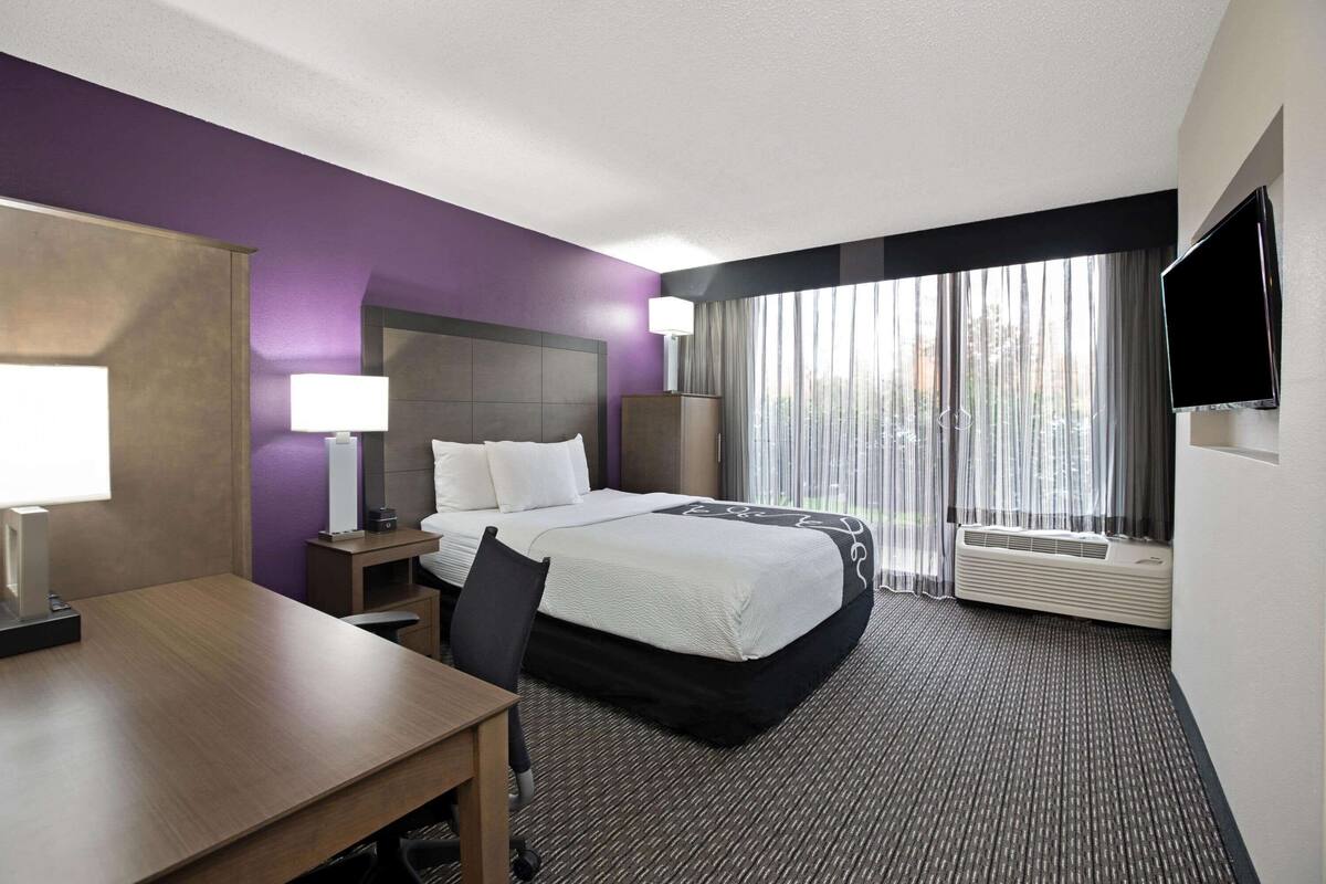 A hotel room with a bed, desk, chair, lamp, TV, and large window with curtains.