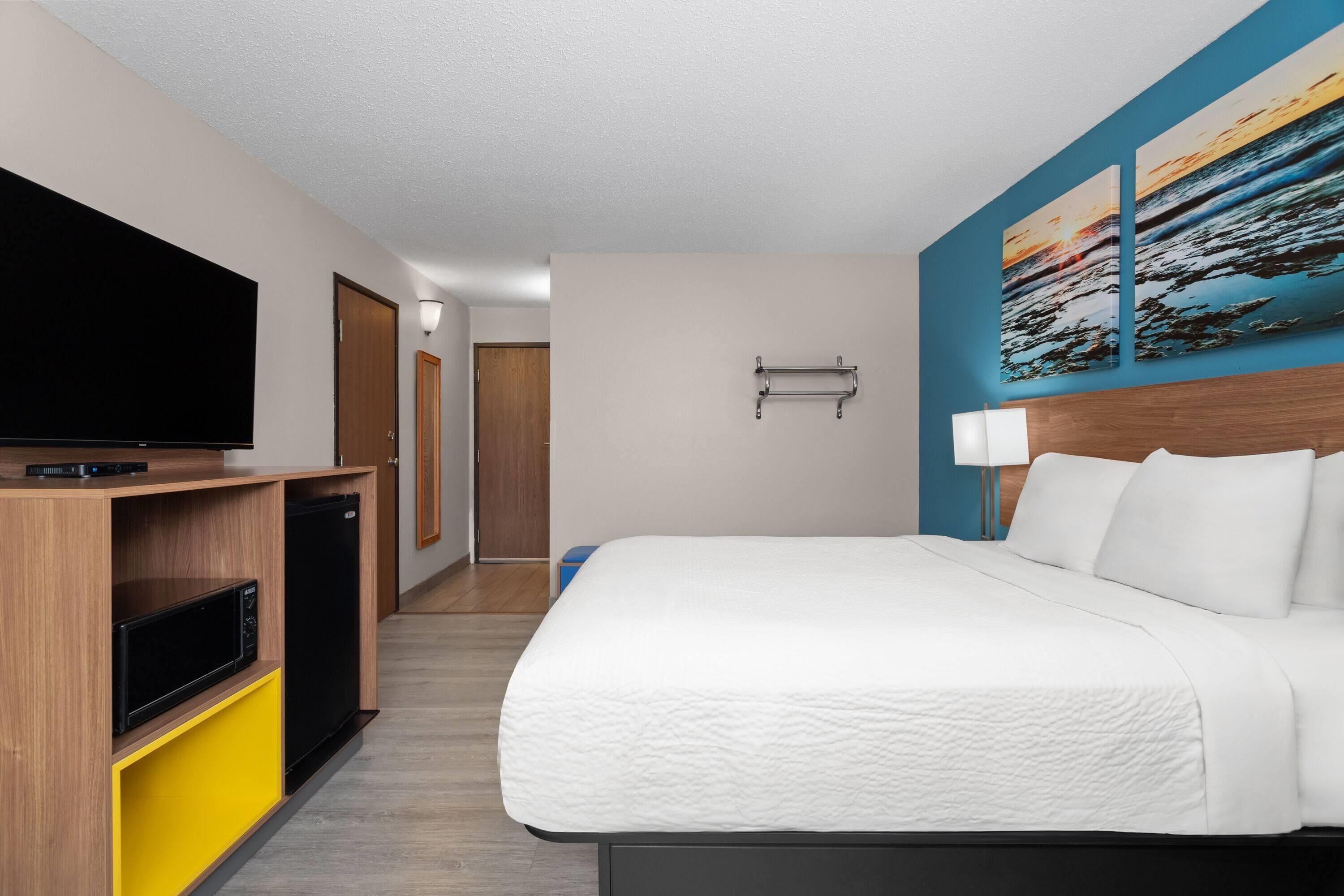 A hotel room with a bed, a TV, a small kitchenette, and a hallway leading to another room.
