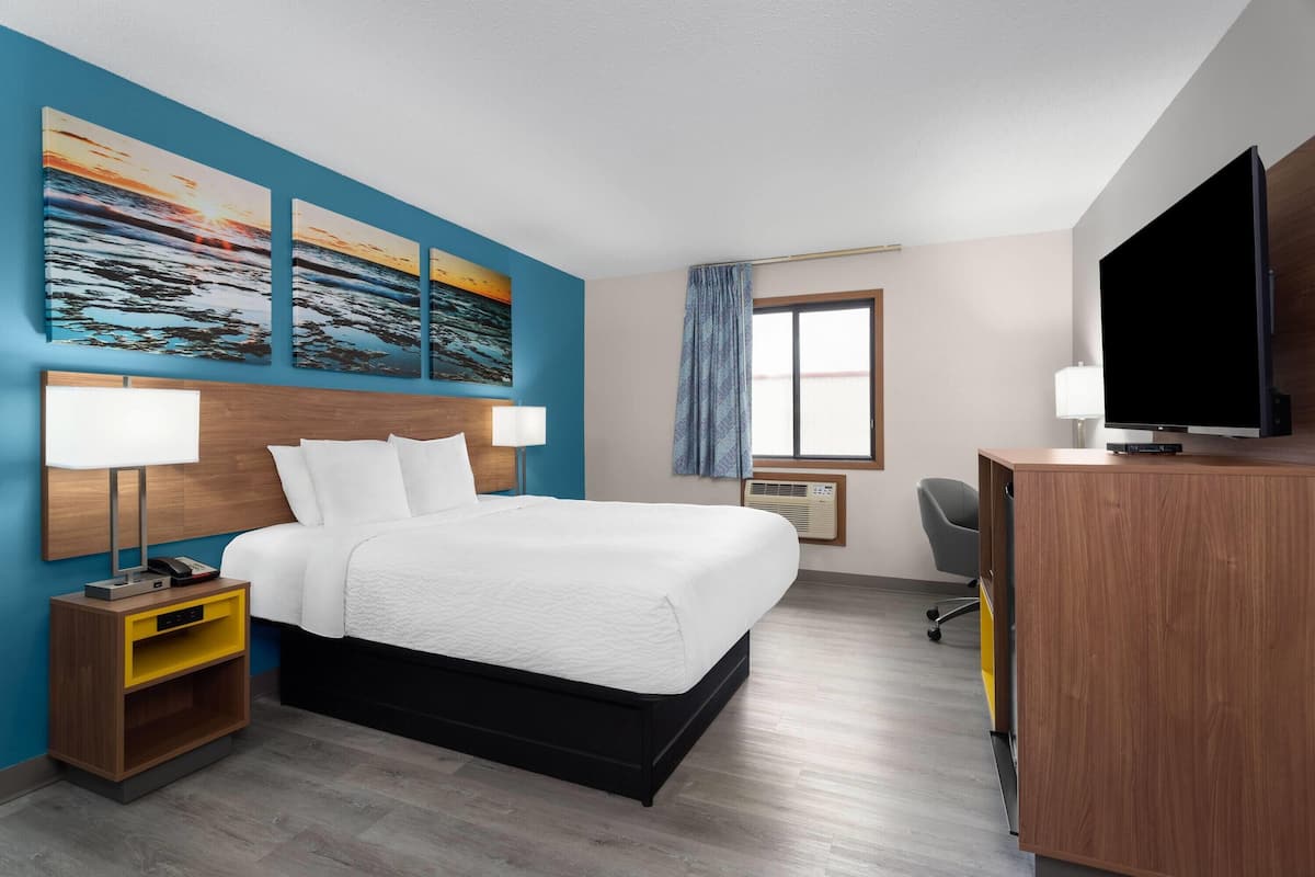 A hotel room with a bed, bedside tables, a desk, a television, and artwork on the wall.