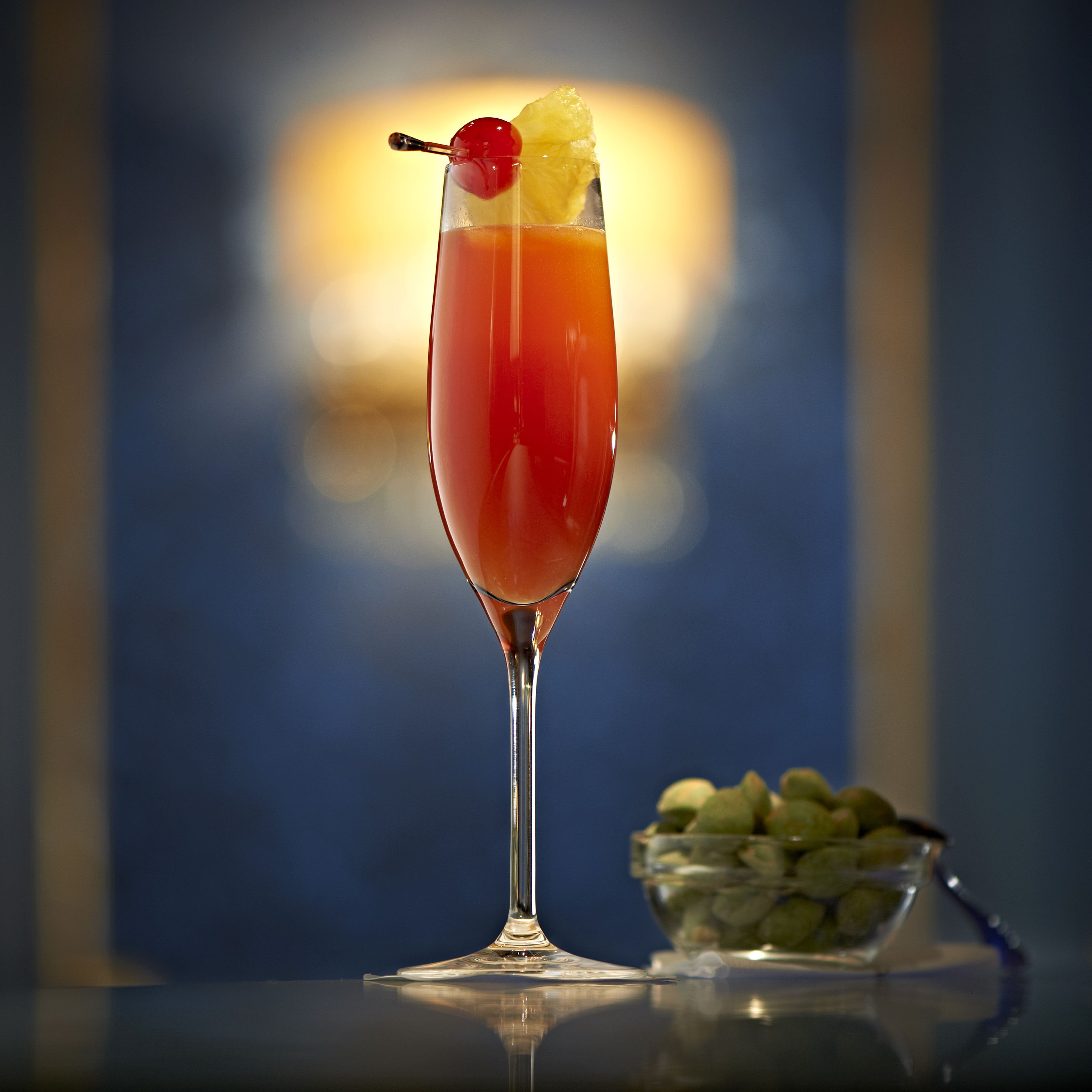 Un cocktail servi dans un verre à pied, décoré d’une cerise et d’un morceau d’ananas, posé sur une surface réfléchissante avec un arrière-plan flou.