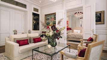 A luxurious room with a white sofa, a glass coffee table, a decorative vase with flowers, a gold-framed armchair, and a bed with a red accent pillow.
