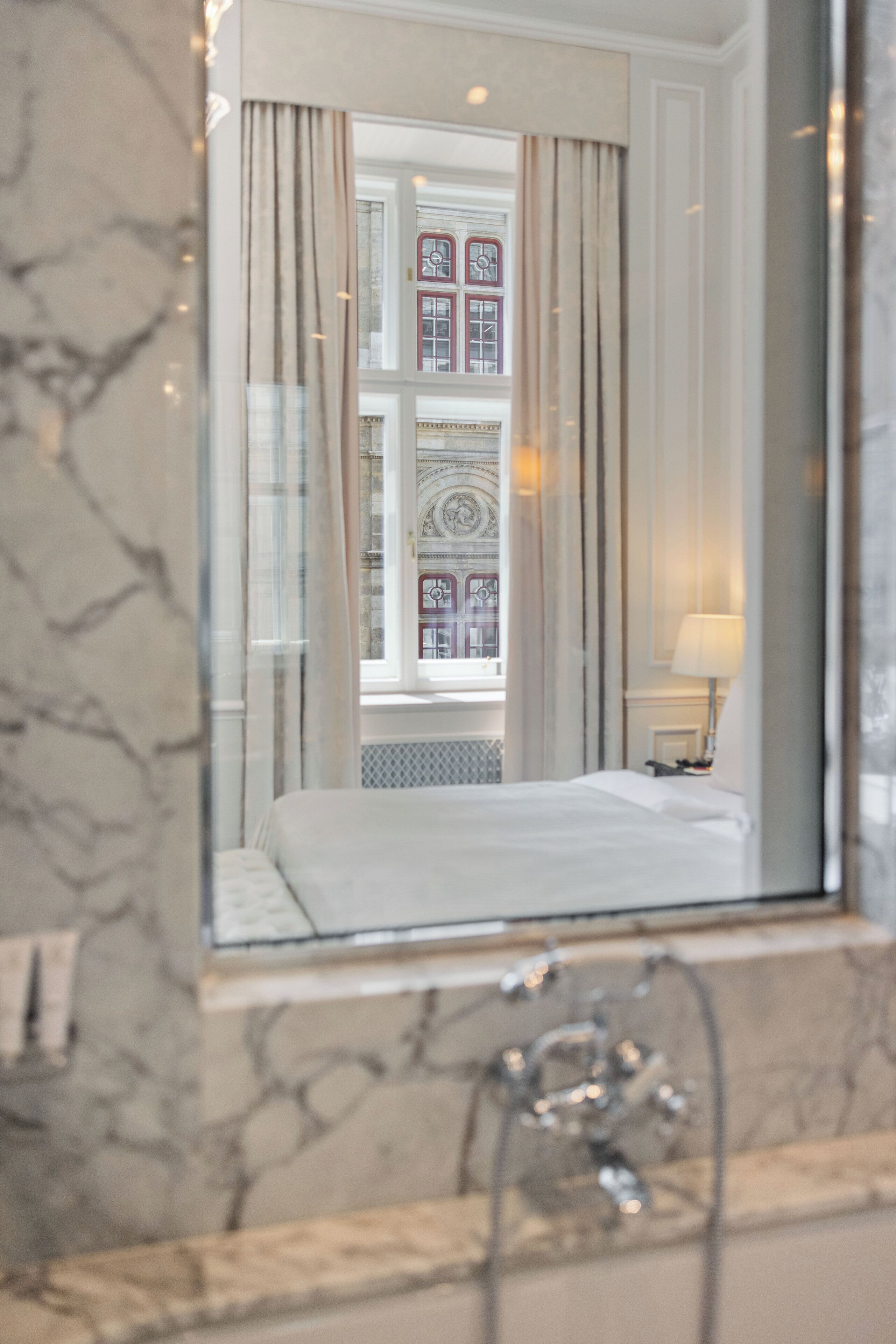 Habitación de hotel con un espejo grande que refleja una ventana con cortinas, una cama y una lámpara en una mesita de noche.