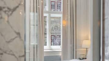 Habitación de hotel con un espejo grande que refleja una ventana con cortinas, una cama y una lámpara en una mesita de noche.