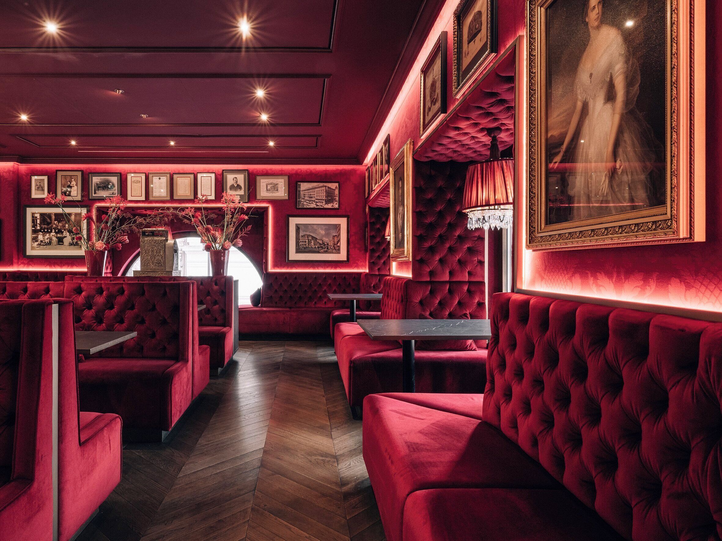 Un restaurante tenuemente iluminado con cabinas y mesas de terciopelo rojo, cuadros en las paredes y un candelabro.