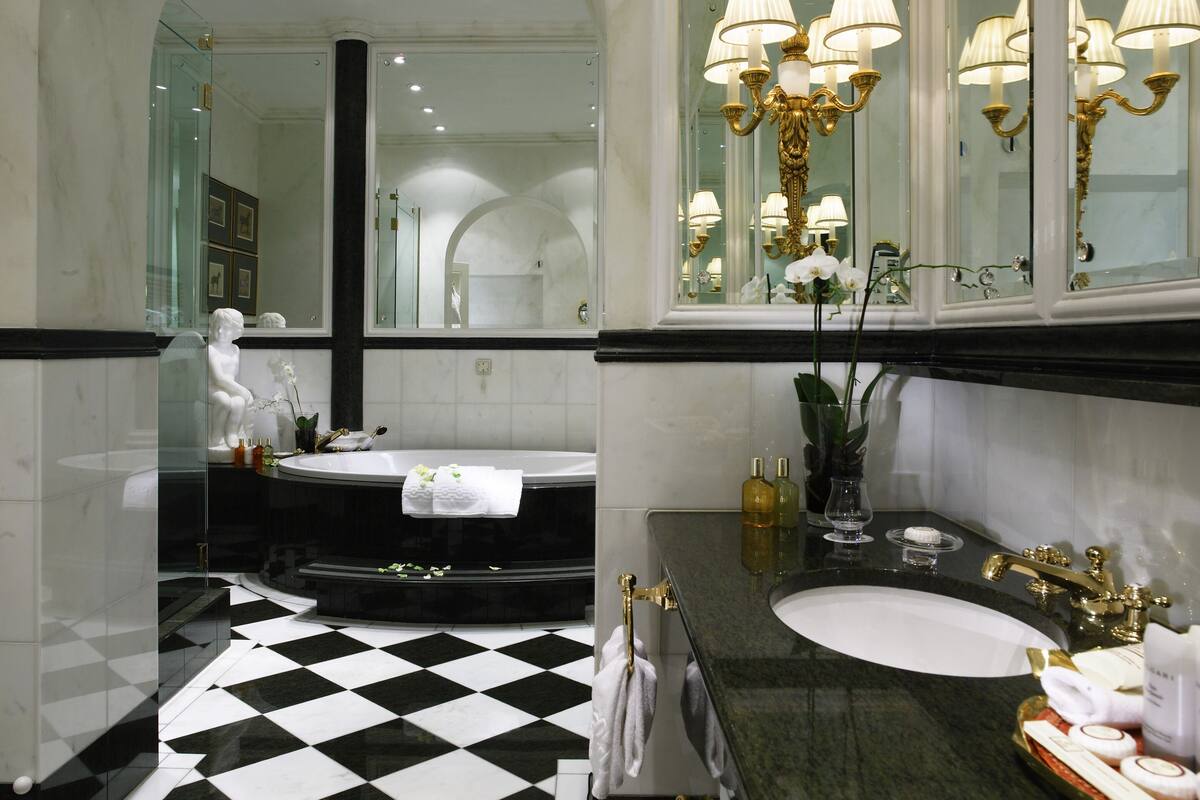 Ein luxuriöses Badezimmer mit einem schwarz-weißen Schachbrettfußboden, einer freistehenden Badewanne, einem großen Spiegel mit einer Chandelier und einem Waschtisch mit einem goldenen Wasserhahn.