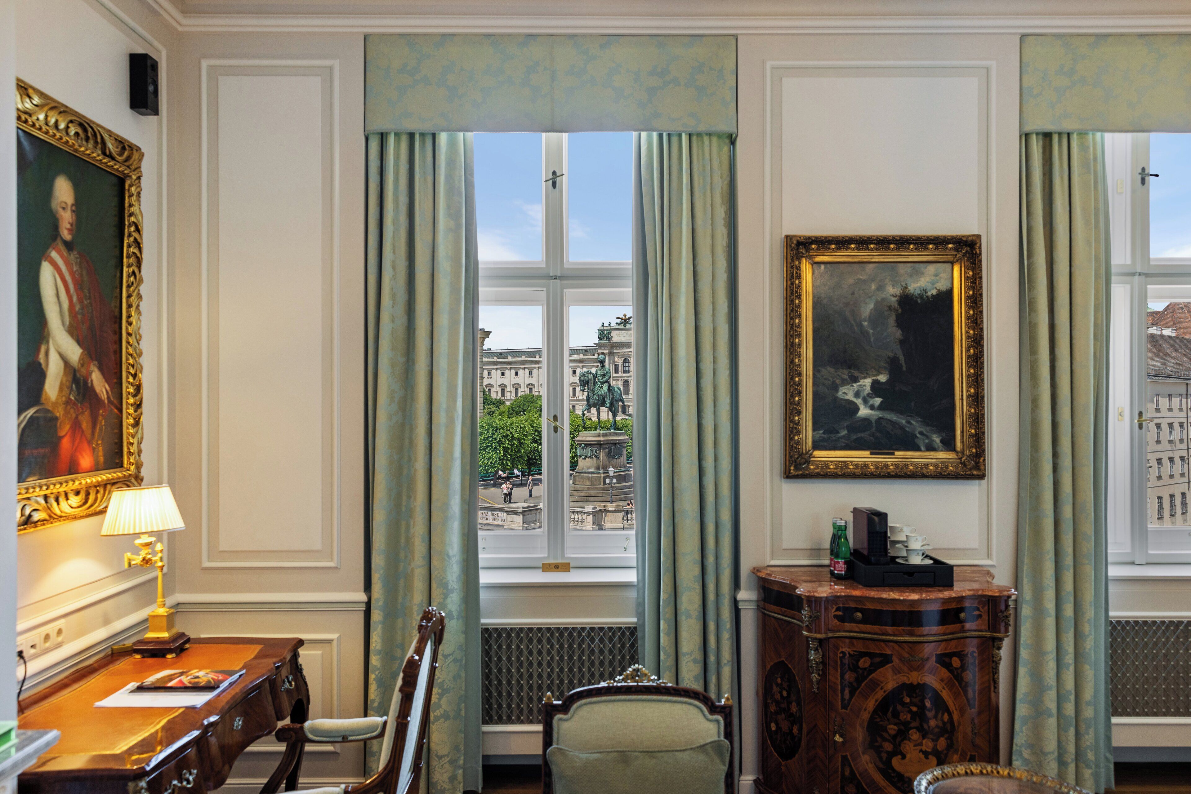 Quarto com escrivaninha, cadeira e vista para uma fonte e edifícios.