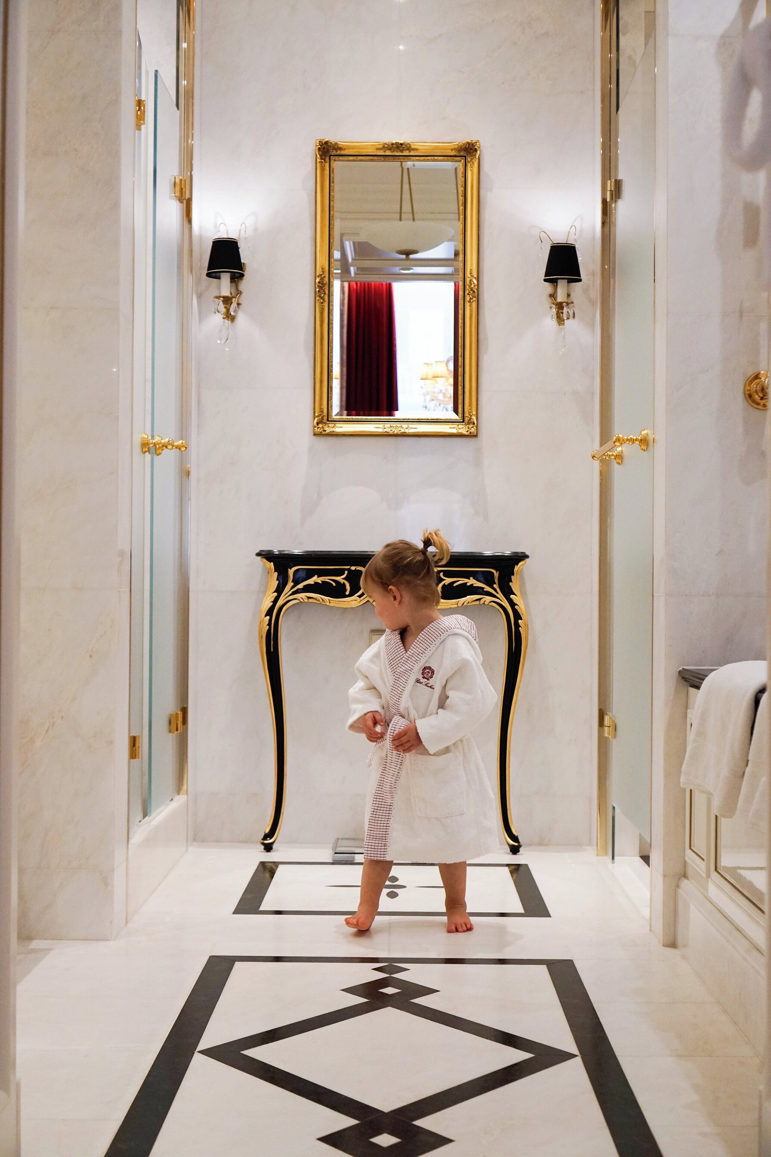 A child in a robe standing in a hallway with a marble floor, a gold-framed mirror, and wall sconces.
