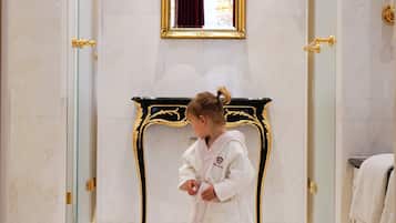 A child in a robe standing in a hallway with a marble floor, a gold-framed mirror, and wall sconces.