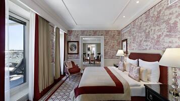 Habitación de hotel con una cama grande, un área de estar con una silla, vista por una ventana y una puerta que da a un comedor.