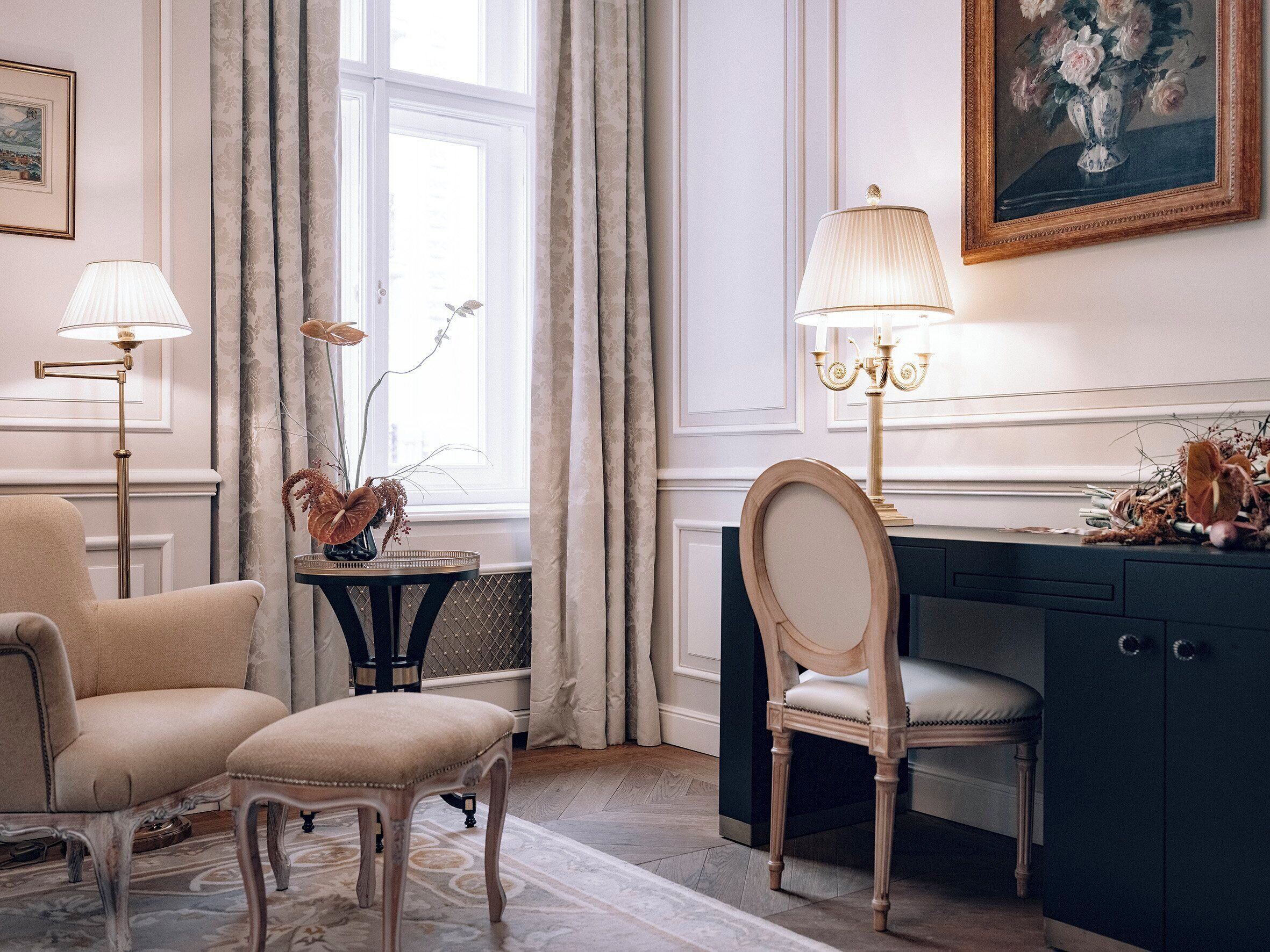 A room with a desk, two chairs, a lamp, a vase with flowers, and a painting on the wall.