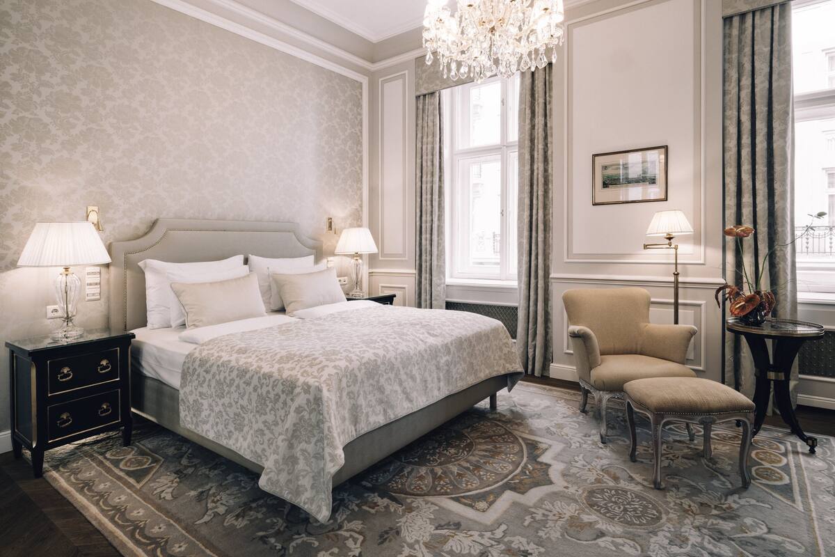 Ein klassisches Hotelzimmer mit einem großen Bett, zwei Nachttischen mit Lampen, einem Sessel, einem kleinen Tisch mit einer Vase und einer Kandelaber.