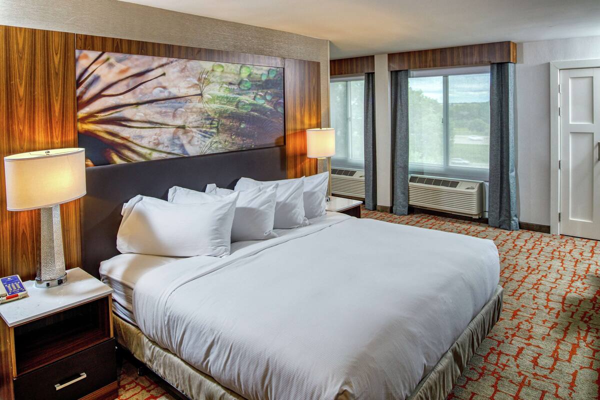 Una habitación de hotel con una cama grande, dos lámparas de noche, una mesita de noche, un aire acondicionado instalado en la pared y una ventana con cortinas.