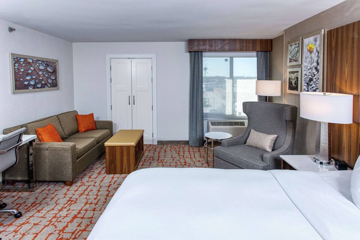Una habitación de hotel con cama, sofá, escritorio, silla, mesita y ventana con cortinas.