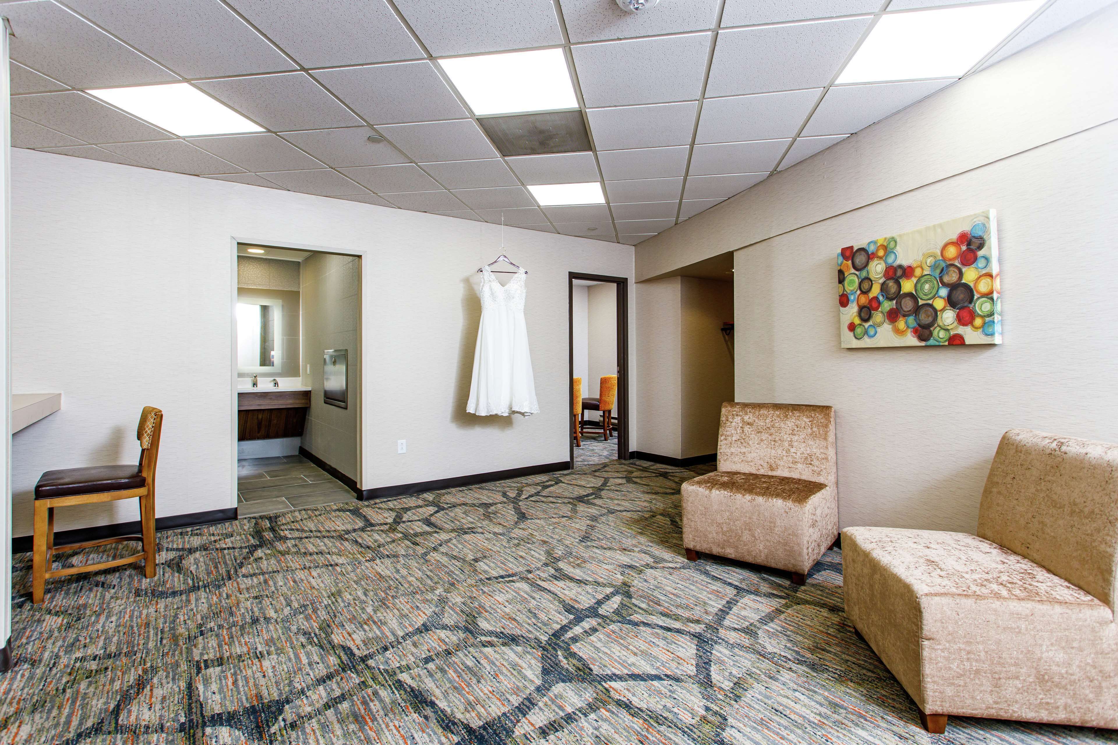 A hotel room with a patterned carpet, a hanging white dress, and a colorful abstract painting on the wall.