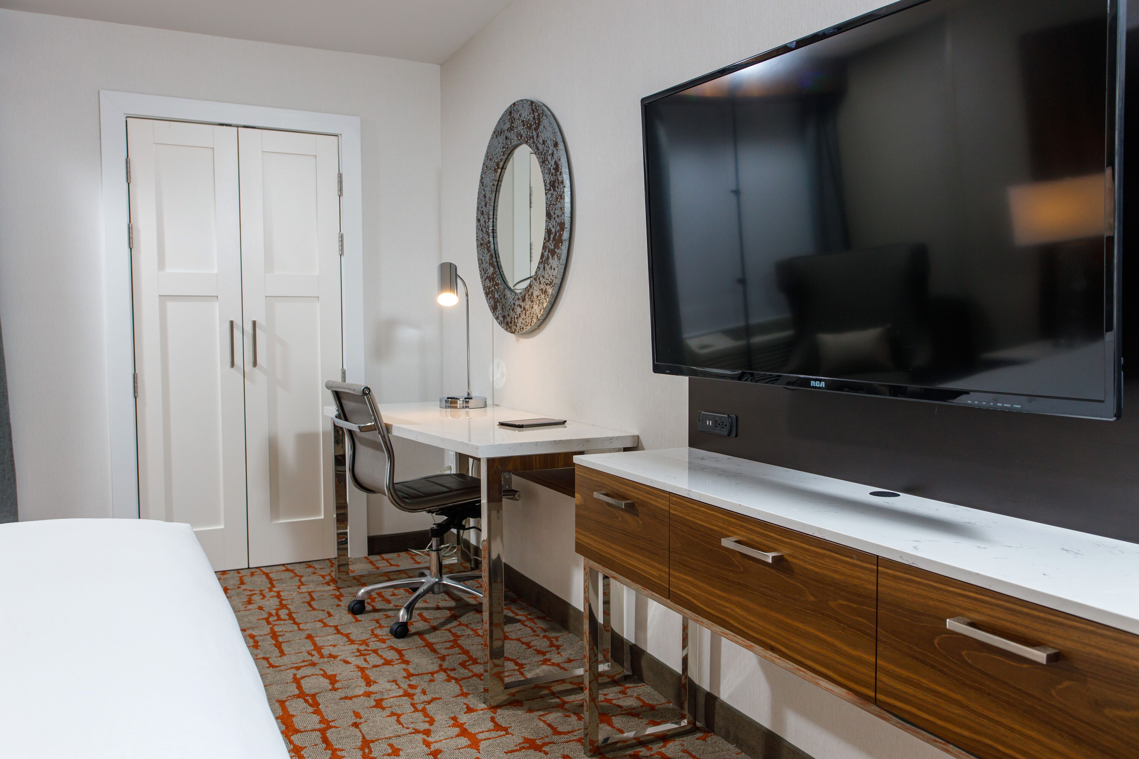 A hotel room with a bed, a desk with a chair, a television, a mirror, and a wardrobe.