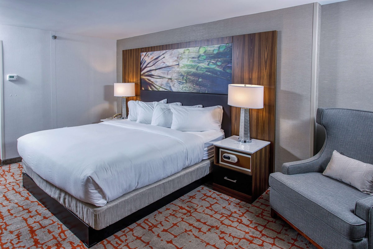 Una habitación de hotel con una cama grande, dos lámparas de noche, una mesita de noche, un sillón gris y una alfombra estampada.