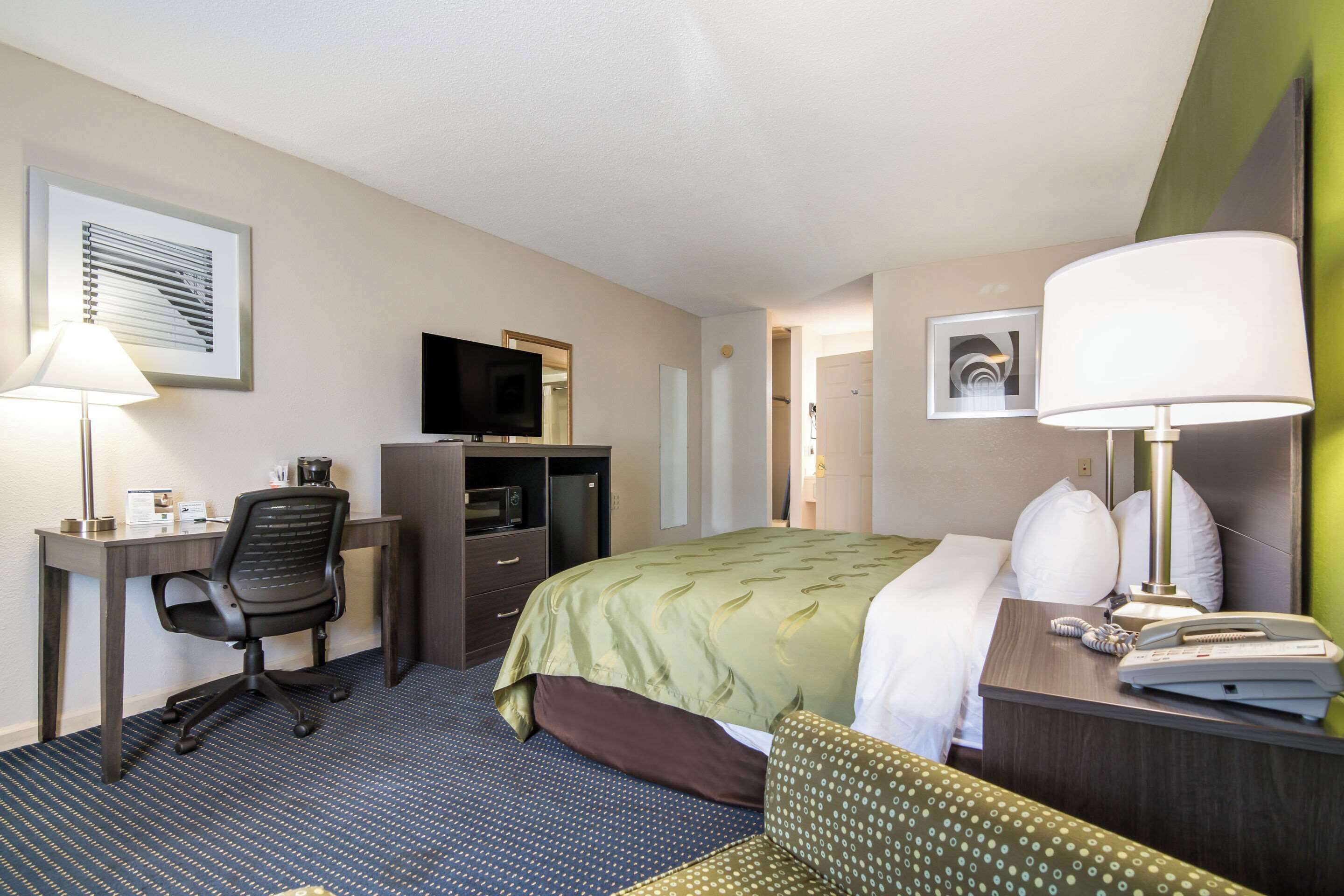 A hotel room with a bed, desk, chair, TV, and a lamp.