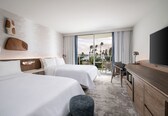 A hotel room with two beds, a desk, a TV, and a balcony with a view of palm trees.