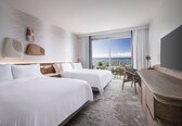 A hotel room with two beds, a large window with a view of the ocean, a desk with a chair, and a flat-screen TV.
