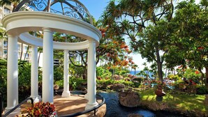 A gazebo with white columns and a metal roof, surrounded by lush tropical plants and a small water feature.