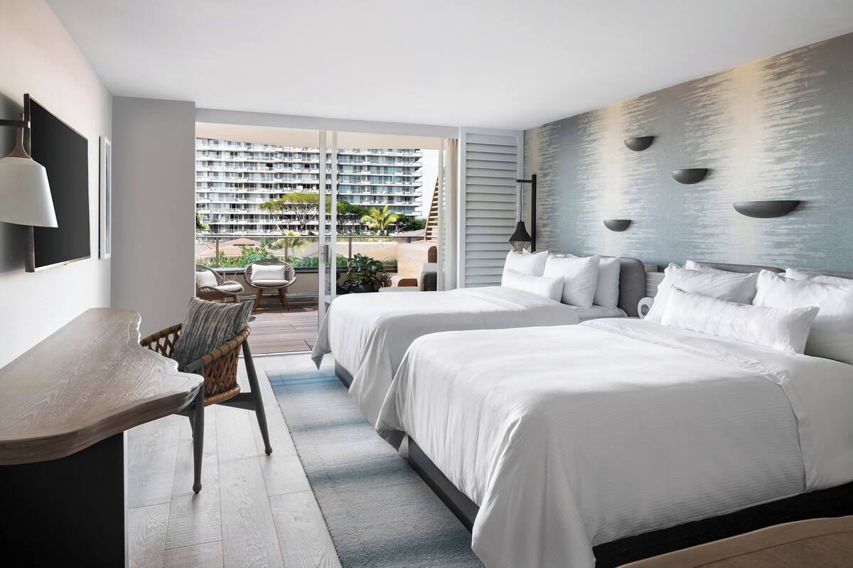 Habitación de hotel con dos camas, un escritorio y vistas a un edificio alto y zona exterior.