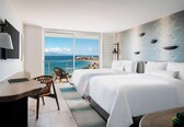 A hotel room with two beds, a desk, and a chair. There is a large window offering a view of the ocean and a distant shoreline.