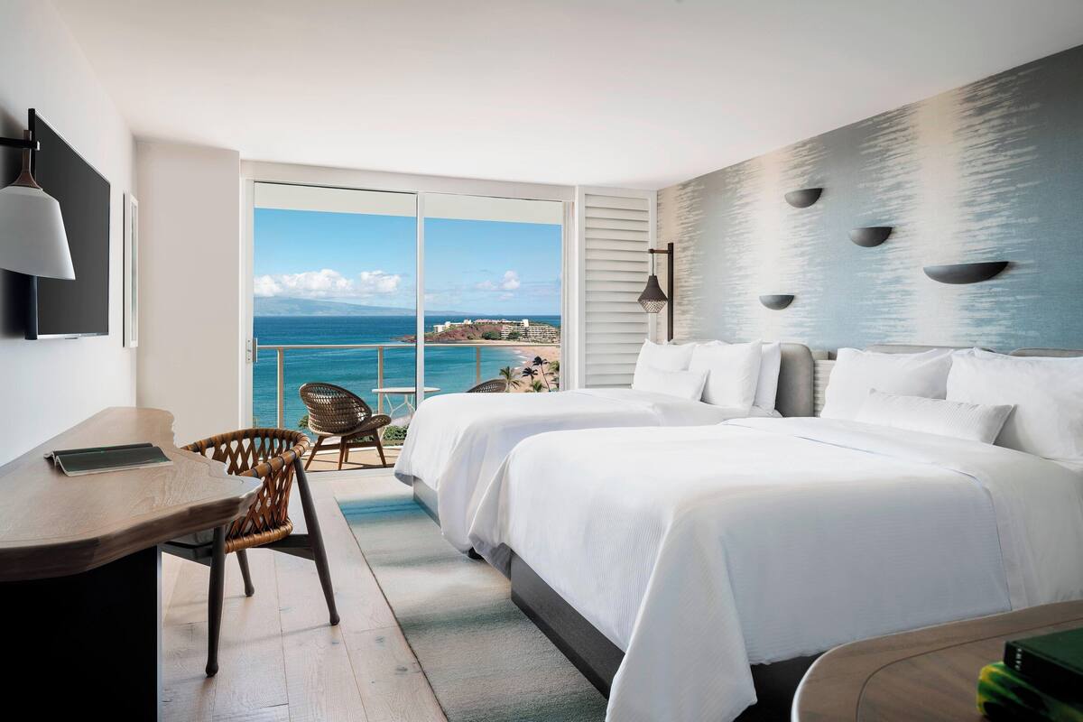 Habitación de hotel con dos camas, un escritorio y una silla. Amplia ventana con vista al océano y a la costa distante.
