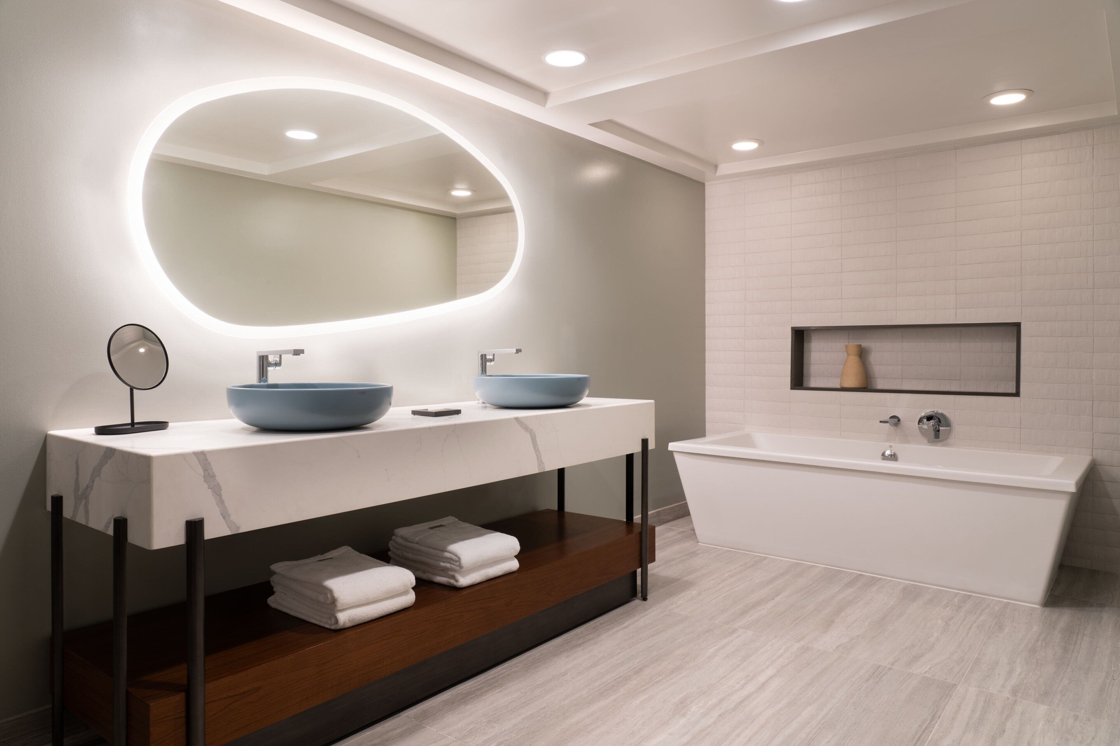 Un baño moderno con un espejo ovalado grande, dos lavamanos, una bañera y un estante con toallas.