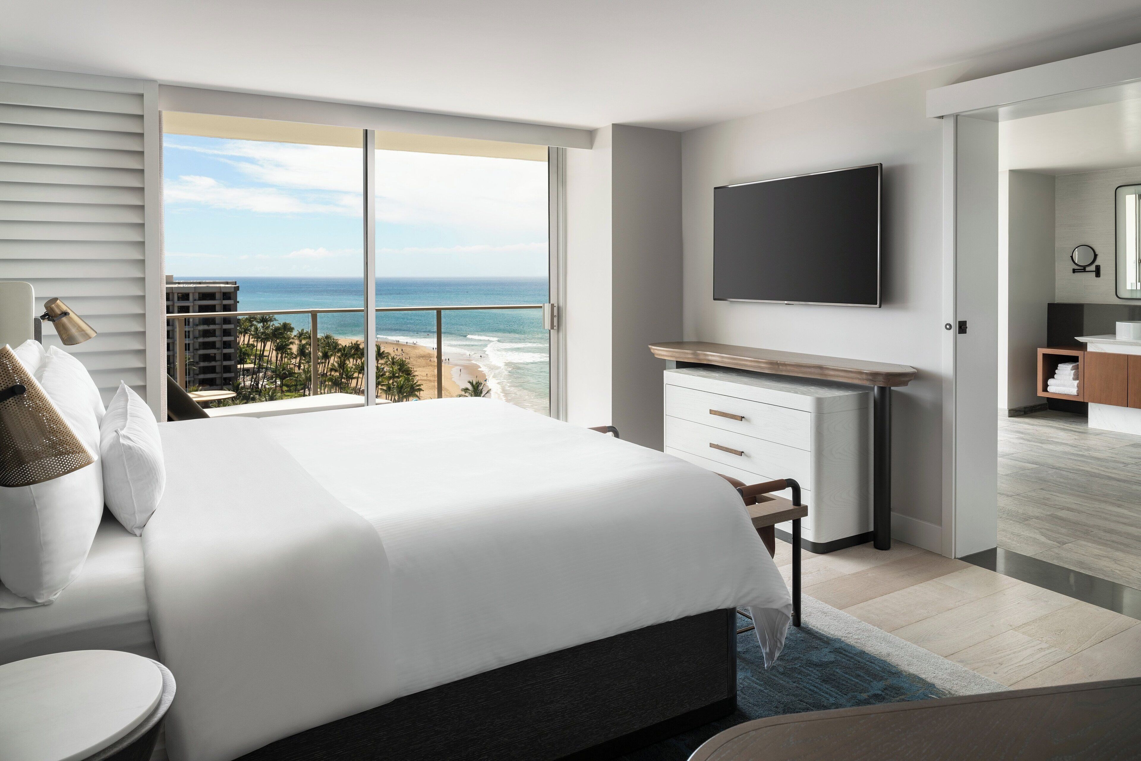 Habitación de hotel moderna con una cama grande, un escritorio, un televisor de pantalla plana y un balcón con vistas a la playa y al océano.