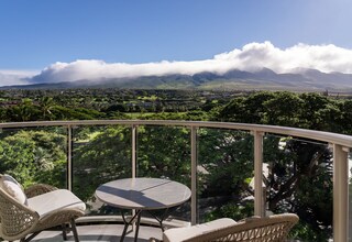 A balcony with wicker furniture and a table, overlooking a lush landscape with mountains and clouds.