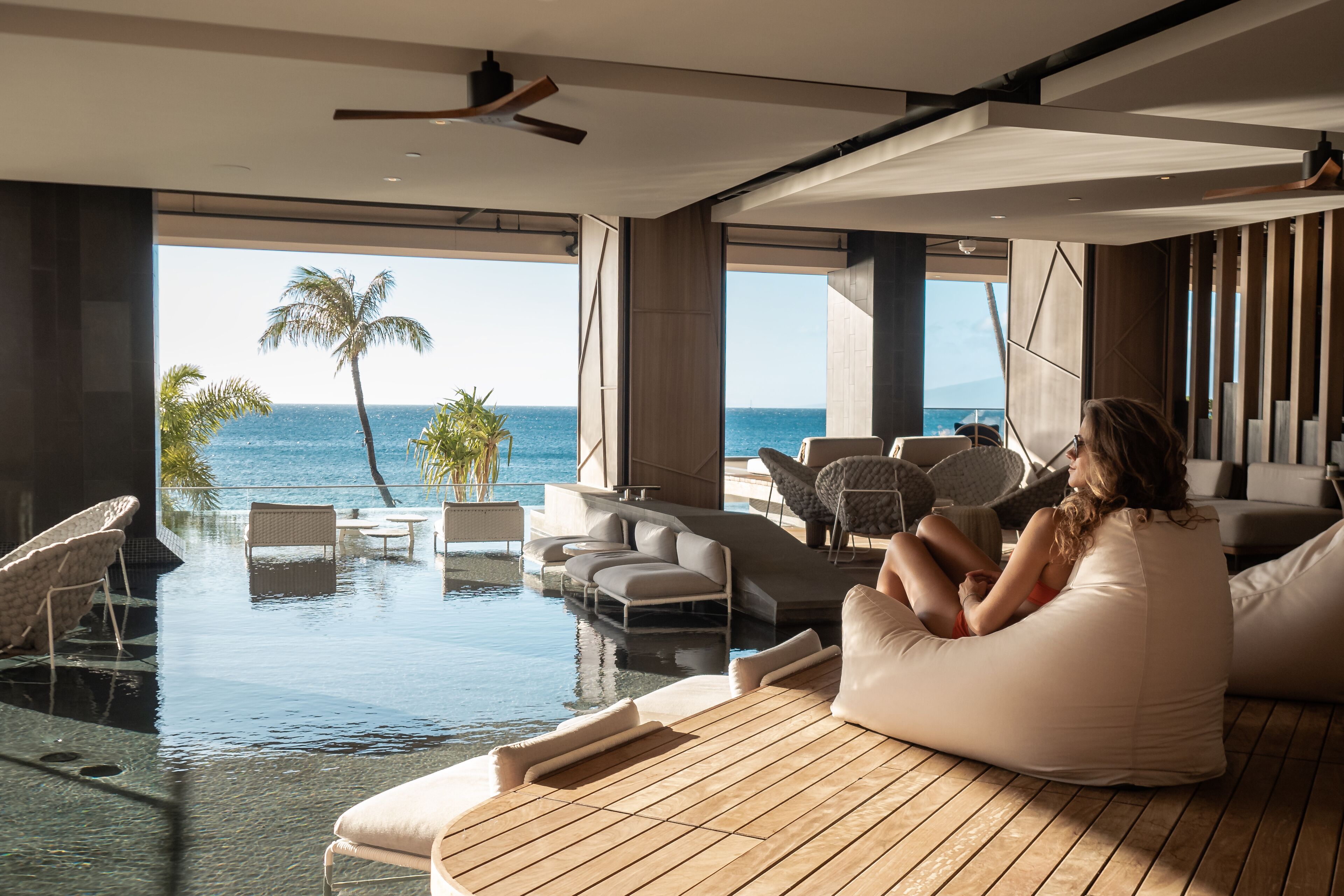 A modern interior with a large window overlooking a pool and the ocean. There are lounge chairs and a sofa.