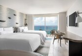 A hotel room with two beds, a desk, a chair, a large window with a view of the sea, and a wall-mounted TV.
