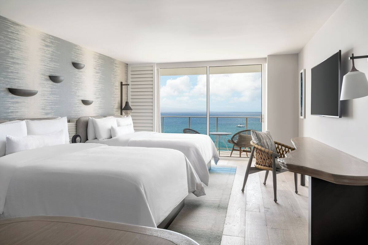 Habitación de hotel con dos camas, un escritorio, una silla, un ventanal con vista al mar y un televisor montado en la pared.