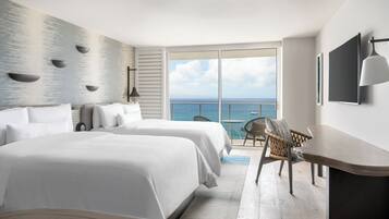 Une chambre d’hôtel avec deux lits, un bureau, une chaise, une grande fenêtre donnant sur la mer et un téléviseur fixé au mur.