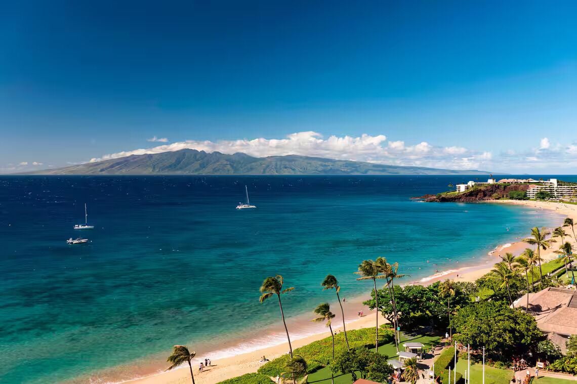 A coastal view with a beach, clear blue water, and a mountain in the distance.