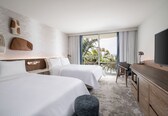 A hotel room with two beds, a TV, a desk, and a balcony with a view of palm trees.