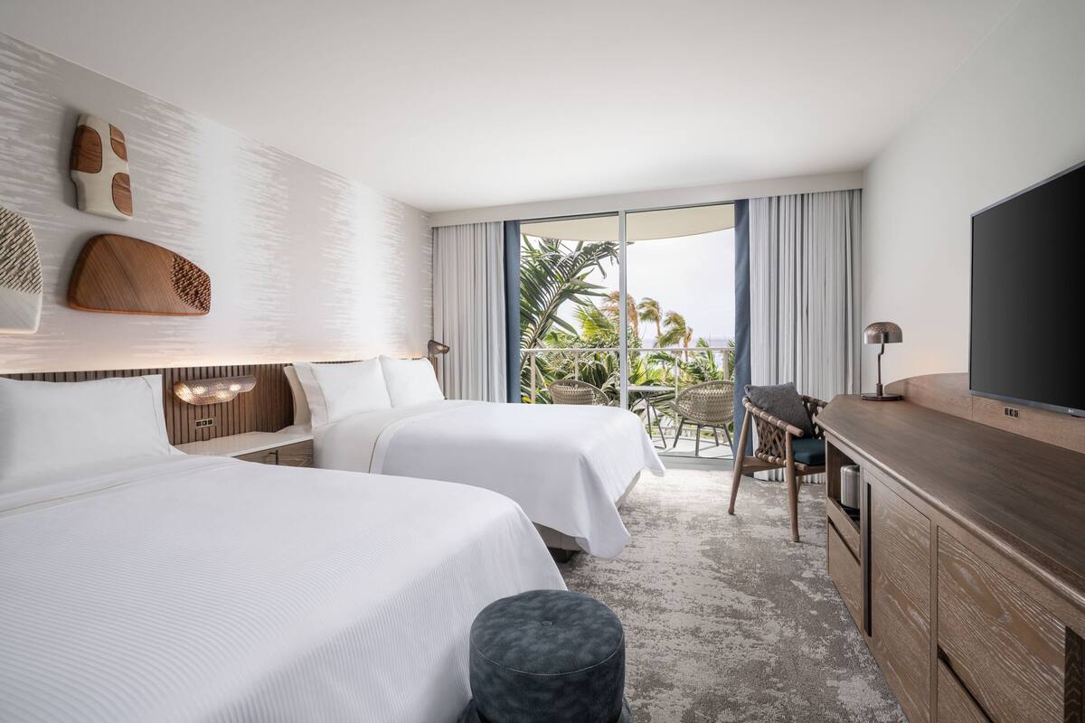 Habitación de hotel con dos camas, televisión, escritorio y balcón con vista a palmeras.