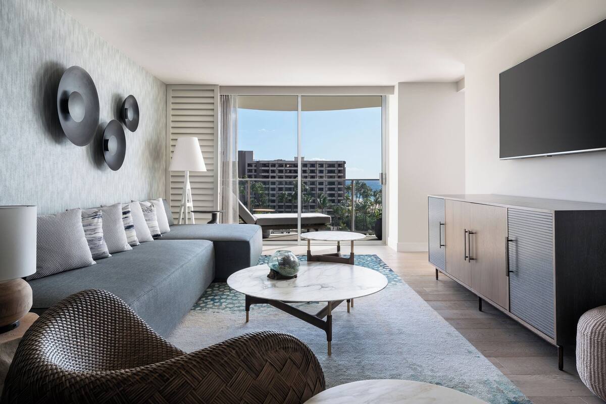 Una sala de estar moderna con un sofá gris, una mesa de centro redonda, sillas de mimbre, un mueble aparador y un televisor grande en la pared.
