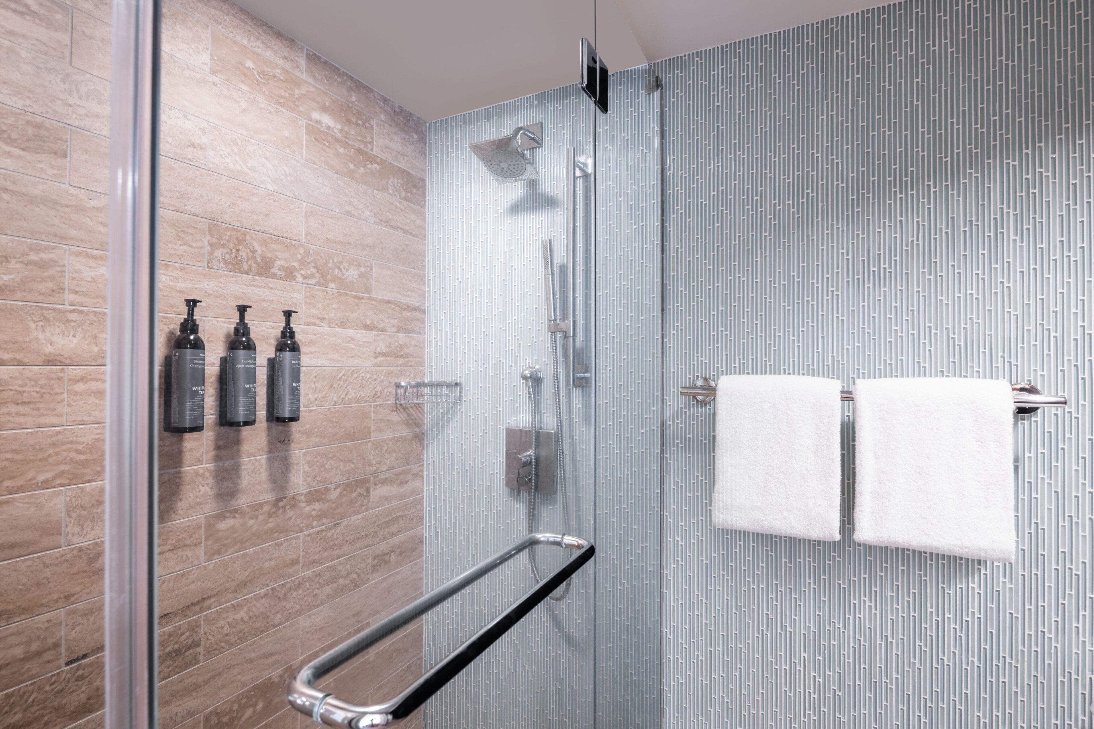 A modern bathroom with a glass shower enclosure, a handrail, and two white towels hanging on a towel rack.