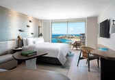 A modern hotel room with a large bed, a sofa, a dining table, and a view of the ocean.