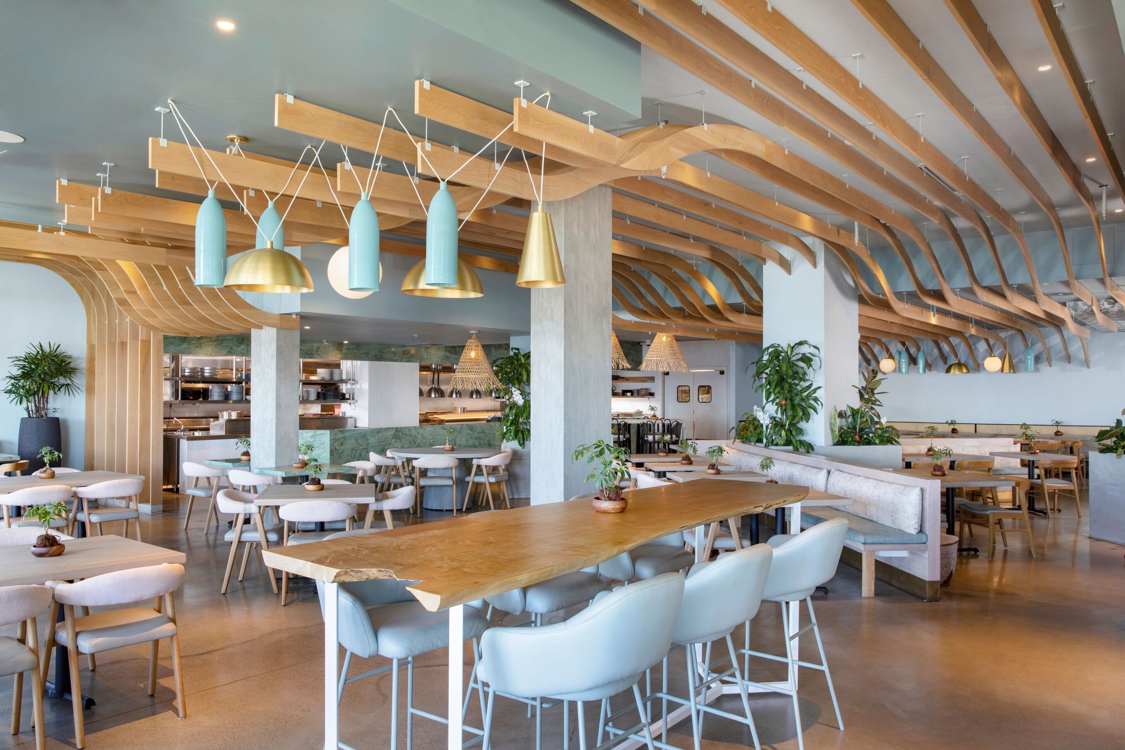 A spacious restaurant with high wooden ceilings, modern pendant lighting, and a variety of seating options including tables, chairs, and a long communal table.