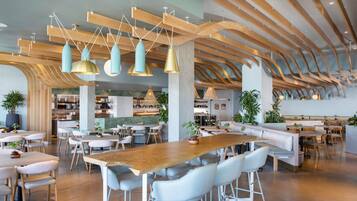 A spacious restaurant with high wooden ceilings, modern pendant lighting, and a variety of seating options including tables, chairs, and a long communal table.