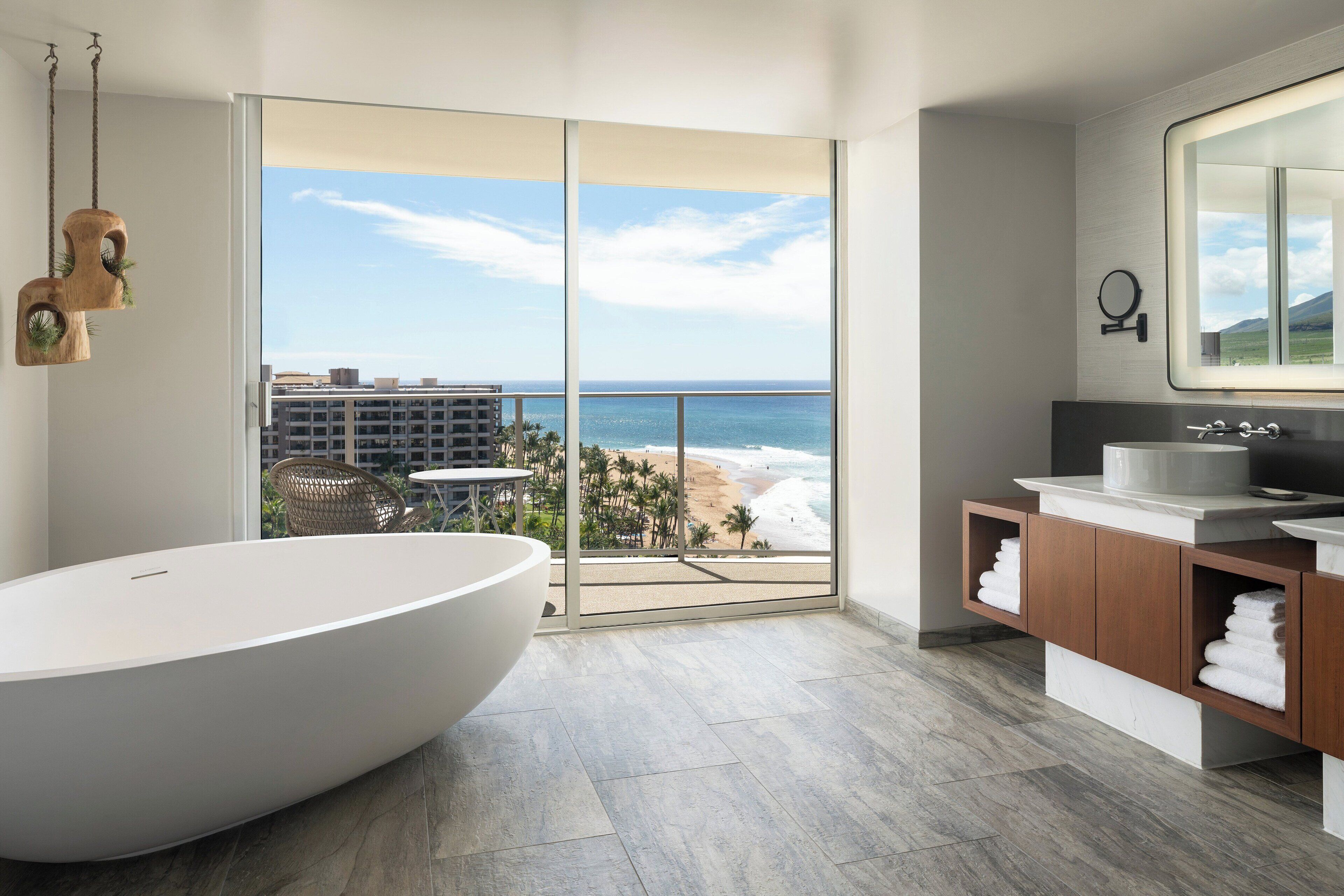 A modern bathroom with a large bathtub, a vanity with a mirror, and a view of the beach and ocean through large sliding doors.