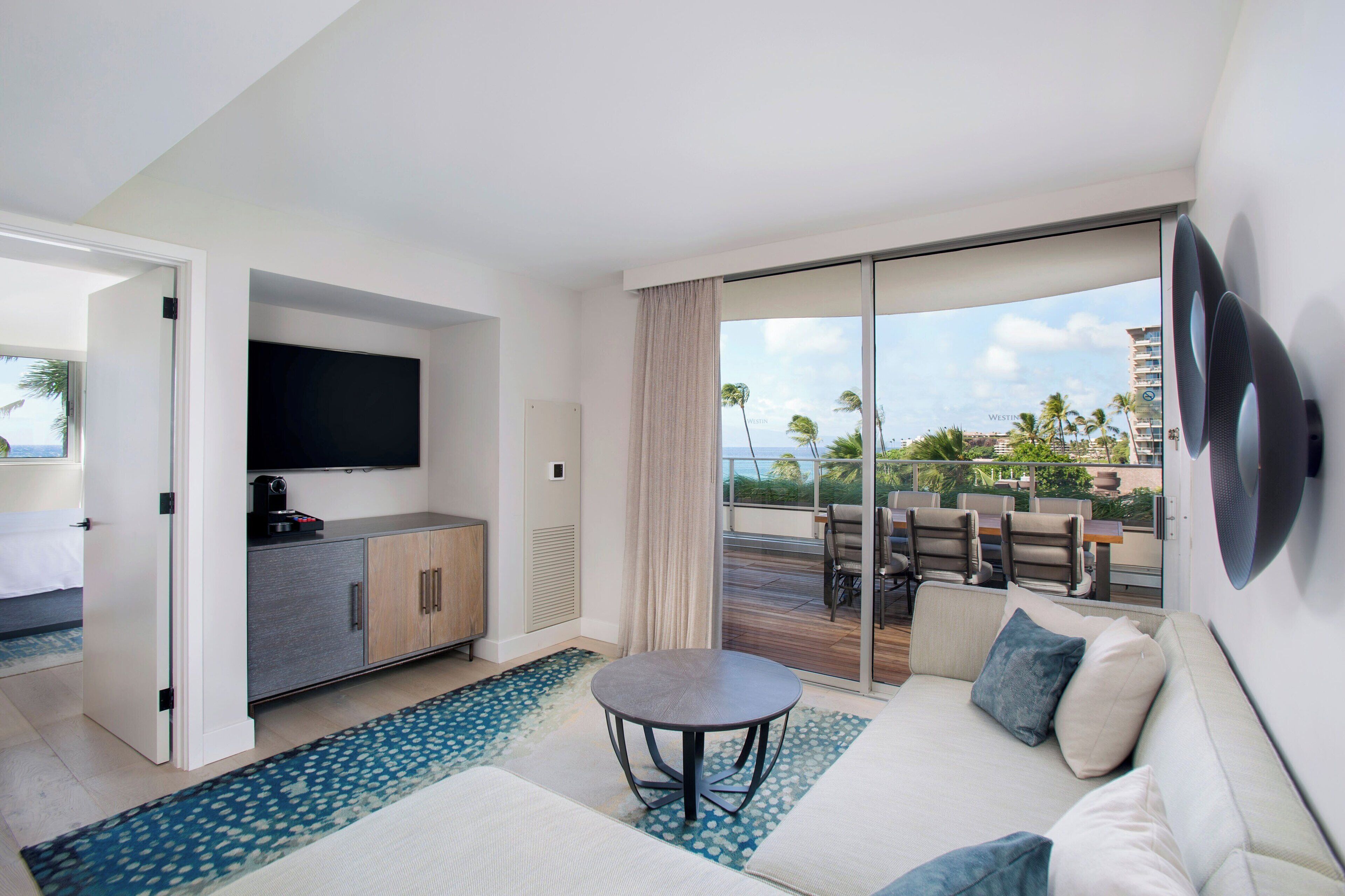 A modern hotel room with a sofa, a flat-screen TV, a dining table, and a balcony with outdoor seating.