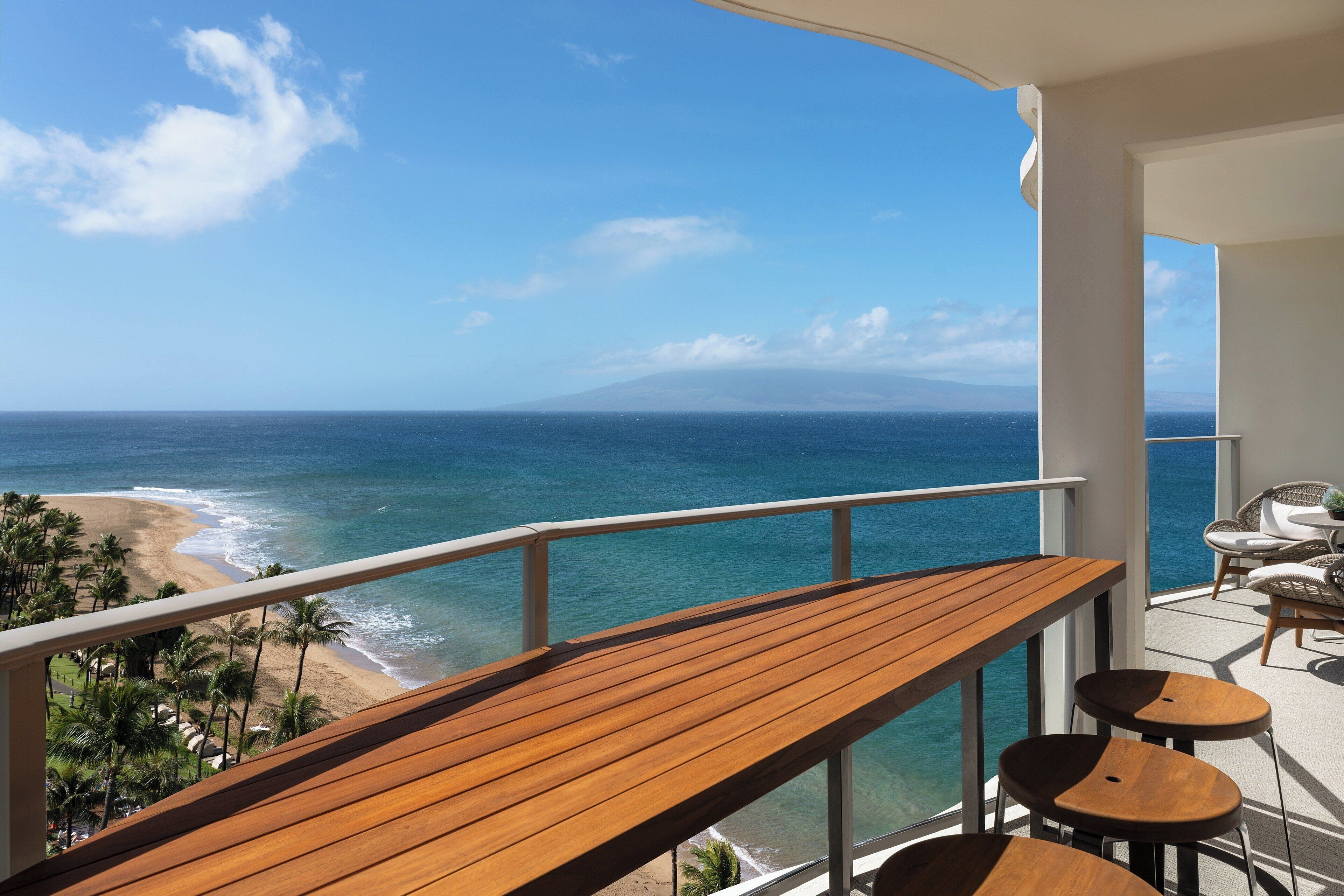 Een balkon met een houten tafel en stoelen, met uitzicht op een strand en de oceaan.