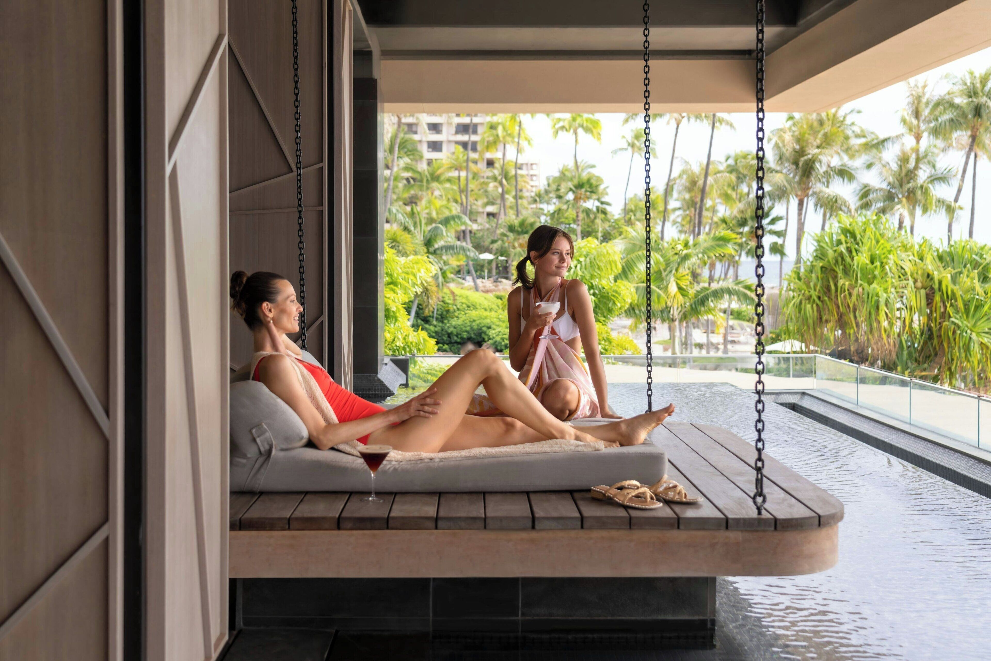 A couple relaxing on a floating lounge chair by a poolside, with a view of palm trees and a building in the background.