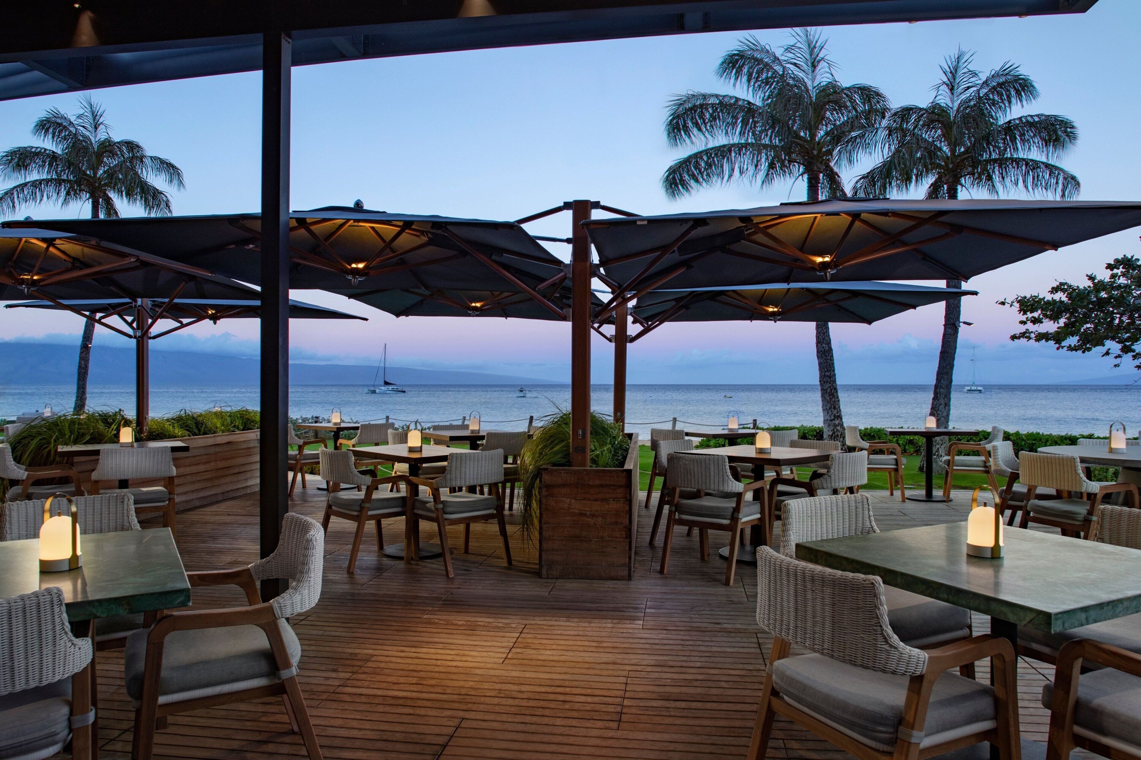An outdoor dining area with tables and chairs, umbrellas, and a view of the ocean.