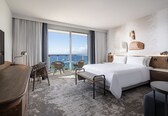 A hotel room with a large bed, a desk, a chair, a small table, and a balcony with a view of the sea.