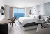 A modern hotel room with a large bed, a dining table, a chair, and a view of the ocean.